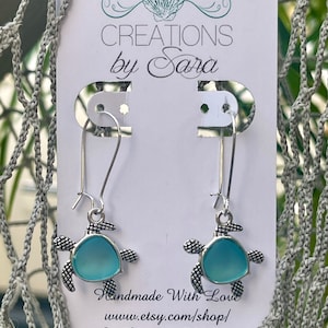 Peut inclure: Une paire de boucles d'oreilles en forme de tortue argentées avec des accents en verre bleu clair. Les boucles d'oreilles sont présentées sur une carte blanche avec le texte "CREATIONS by Sara" et "Handmade With Love www.etsy.com/shop/ Sea Creations By Sara".