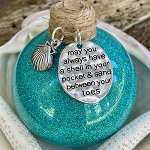 Könnte beinhalten: Türkiser Glitzer-Weihnachtsbaumschmuck mit einem silbernen Anhänger, auf dem steht "Möge du immer eine Muschel in deiner Tasche und Sand zwischen deinen Zehen haben".
