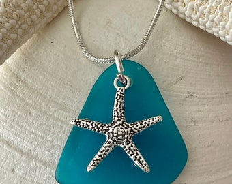 May include: A teal sea glass pendant necklace with a silver starfish charm. The pendant hangs from a delicate silver chain. The necklace is displayed on a white seashell, evoking a beachy, coastal theme.