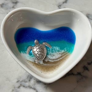 May include: A heart-shaped white ceramic dish with a beach scene design. The interior features blue and turquoise resin representing the ocean, with a silver-colored turtle charm. The dish is designed for small items.