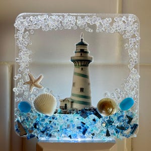 May include: A decorative night light with a lighthouse design. The rectangular light has a clear border with small, clear beads. The lighthouse is white with blue stripes, and the base has seashells and blue glass pieces.