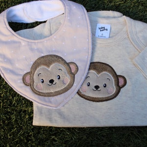 May include: A beige baby bodysuit with a brown monkey embroidered on the front. The bodysuit has a white label that says "baby berry" and "0-6 months". A beige bib with white polka dots and a brown monkey embroidered on the front is also included.