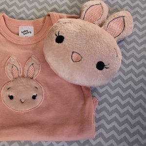 Puede incluir: Un body de bebé rosa con un conejito bordado en rosa en la parte delantera. El body tiene una etiqueta que dice "baby berry".