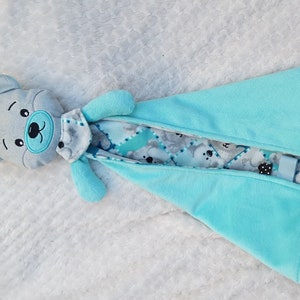 Puede incluir: Una manta de seguridad para bebé suave, azul y gris con un oso de peluche gris adjunto. La manta está hecha de una tela suave y afelpada y presenta un patrón de osos grises sobre un fondo azul y blanco.