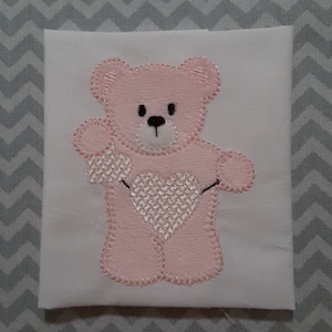 May include: Pink embroidered teddy bear holding a heart on a white fabric background.