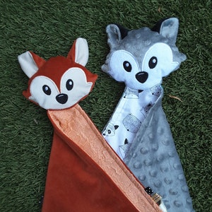 May include: Two plush animal security blankets, one with a fox head and the other with a wolf head. The fox blanket is orange and the wolf blanket is gray. Both blankets have a white and black patterned fabric on the back.