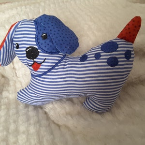 Op de afbeelding: Een stoffen hondenspeelgoed met blauw-witte gestreepte stof. De hond heeft blauwe oren met stippen, geborduurde blauwe vlekken en een rode tong. De staart is rood met witte stippen. Het speelgoed ligt op een witte, zachte ondergrond.