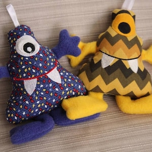 Pode incluir: Dois brinquedos de pelúcia com a forma de monstros. Um é azul com dentes brancos e um único olho. O outro é amarelo com listras pretas e marrons e dentes brancos.