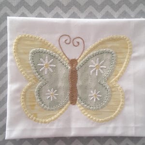 May include: A white fabric with a yellow and green embroidered butterfly with brown accents and white flower details.