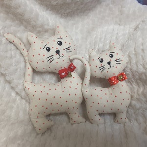 Può includere: Due giocattoli per gatti in tessuto bianco con pois rossi e fiocchi rossi. Il gatto più grande ha un campanello sul suo fiocco.
