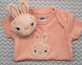 May include: A peach-colored baby onesie with a brown embroidered bunny on the front. The onesie has a white tag that says "baby berry".