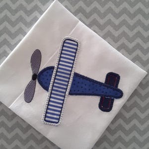 May include: A blue and white airplane applique with a gray propeller. The applique is sewn onto a white fabric background.