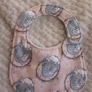May include: A pink baby bib with a white trim and a repeating pattern of gray elephants with stars. The elephants are wearing a white hat.