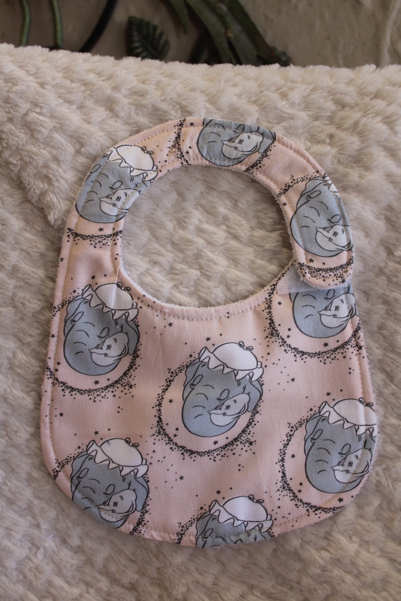 May include: A pink baby bib with a white trim and a repeating pattern of gray elephants with stars. The elephants are wearing a white hat.