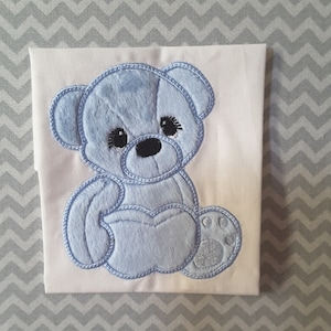 May include: A light blue embroidered teddy bear design on a white fabric background.
