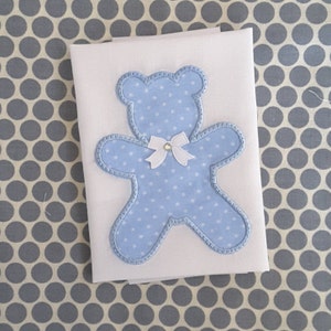 Puede incluir: Una aplicación de oso de peluche azul con lunares blancos y un lazo blanco. La aplicación está sobre un fondo de tela blanca.