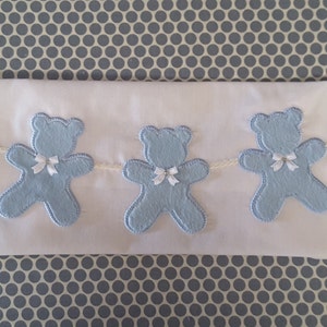 May include: White baby blanket featuring three light blue teddy bear appliqués, each with a white bow. The bears are linked by a cream-coloured string. The background has a blue and white polka dot pattern.