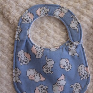 May include: A blue baby bib with a white border and a pattern of cartoon baby elephants with pink ears. The elephants are all smiling and have a light blue background.