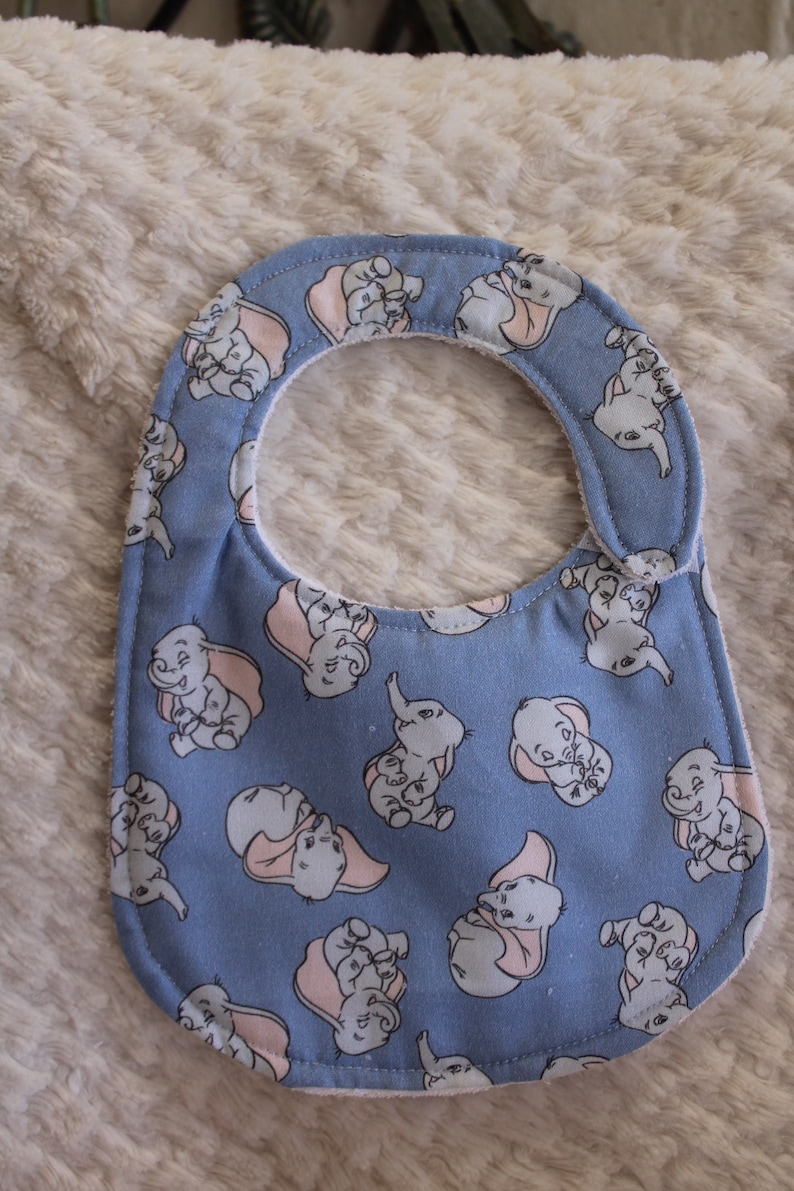 May include: A blue baby bib with a white border and a pattern of cartoon baby elephants with pink ears. The elephants are all smiling and have a light blue background.