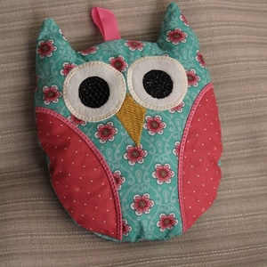 Może przedstawiać: Poduszka w kształcie sowy w kolorze turkusowym z różowymi kropkami i wzorami kwiatowymi. Sowa ma białe filcowe oczy z czarnymi źrenicami i żółty dziób.
