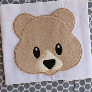 May include: Embroidered brown bear face with black eyes and nose, and a white snout. The bear face is on a white fabric background.