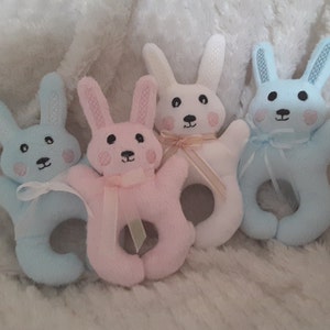 以下が含まれることがあります： 青、ピンク、白のぬいぐるみ製のうさぎのガラガラおもちゃ4個。おもちゃは柔らかい生地で作られており、刺繍された顔をしています。それぞれのおもちゃには、赤ちゃんが握るためのループが付いています。