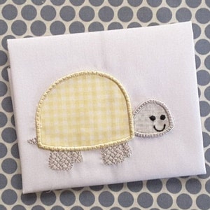 Pode incluir: Uma aplicação de tartaruga em vichy amarelo e branco com uma casca e um rosto bordados em cinza. A tartaruga tem um rosto sorridente com olhos pretos.