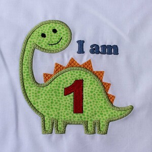 Peut inclure: Applique de dinosaure vert avec un numéro un rouge et le texte "Je suis" en bleu. Le dinosaure a un visage souriant et des pointes orange sur le dos.