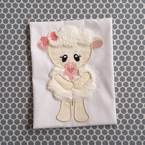 Può includere: Applique ricamata bianca di un agnello che tiene un cuore rosa. L'agnello ha un fiocco rosa sulla testa e sta in piedi su uno sfondo bianco.