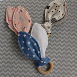 以下が含まれることがあります： 木製の歯固めリングに、布製の飾り付けが3つ付いています。飾り付けは、柔らかくカラフルな布でできており、それぞれ異なる模様をしています。1つの飾り付けは、白い背景に青い水玉模様、もう1つは、ピンクの背景に幾何学模様、そして3つ目は、白い背景に白黒の猫の模様が描かれています。