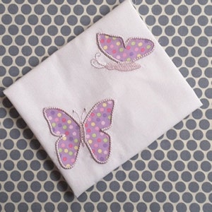 Könnte beinhalten: Weißer Stoff mit zwei gestickten Schmetterlingen. Die Schmetterlinge sind lila mit rosa Umrandungen und haben bunte Punkte.