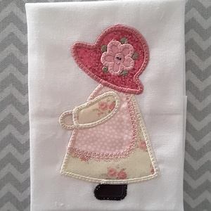 May include: A white tea towel with an embroidered applique of a girl wearing a pink dress and a pink hat with a flower on top.