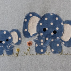 Puede incluir: Una manta de bebé azul con dos elefantes azules bordados con lunares blancos. Los elefantes están de pie en un campo de hierba verde con flores rosas y amarillas.