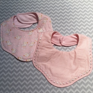 Può includere: Due bavaglini per neonati di colore rosa. Un bavaglino ha un motivo di anatre gialle e bolle bianche su uno sfondo rosa. L'altro bavaglino ha un motivo a pois rosa e bianco con un bordo di perline rosa e bianco.