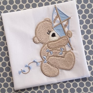 Könnte beinhalten: Eine weiße Babydecke mit einem braunen Teddybär, der einen blauen und weißen Drachen hält. Der Teddybär ist mit blauen Schleifen bestickt.