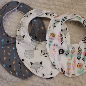 May include: Three baby bibs with different patterns. One bib has a gray background with gold and blue arrows. Another bib has a white background with black and gray foxes and raccoons. The third bib has a white background with pink, gold, and blue feathers.