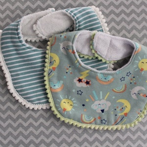 May include: Two baby bibs. One bib has a teal and white striped pattern with white pom-pom trim. The other bib has a light teal background with a colorful celestial pattern, including suns, moons, and clouds, with green pom-pom trim.