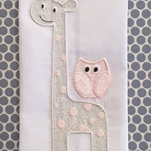 May include: A white fabric with a grey and pink embroidered giraffe and owl. The giraffe has pink polka dots and the owl has a pink body with black eyes.