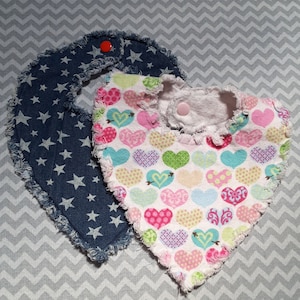 May include: Two baby bibs, one with a blue denim fabric with white stars and the other with a white fabric with a pink, green, and blue heart pattern.