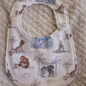 May include: White baby bib with a jungle animal print. The bib features a variety of animals including a zebra, giraffe, kangaroo, and a bear. The animals are all drawn in a cartoon style and are surrounded by palm trees and clouds.