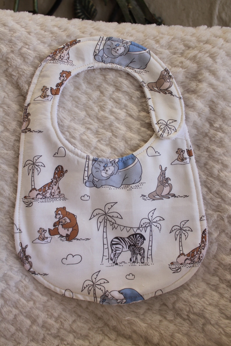 May include: White baby bib with a jungle animal print. The bib features a variety of animals including a zebra, giraffe, kangaroo, and a bear. The animals are all drawn in a cartoon style and are surrounded by palm trees and clouds.