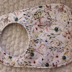 Op de afbeelding: Een witte baby-slabbetje met een bloemen- en konijnenprint. Het slabbetje heeft een zachte, absorberende voering en een veilige sluiting.
