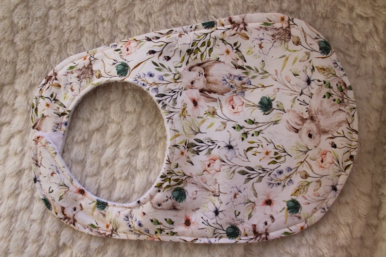 Op de afbeelding: Een witte baby-slabbetje met een bloemen- en konijnenprint. Het slabbetje heeft een zachte, absorberende voering en een veilige sluiting.