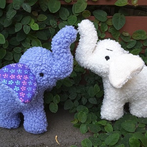 Puede incluir: Dos elefantes de peluche, uno azul con un patrón floral morado y rosa en la espalda y otro blanco. Los elefantes están de pie sobre un fondo de hojas verdes.