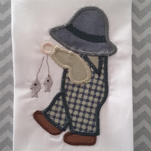 Puede incluir: Una aplicación de fieltro de un niño que lleva una camisa a cuadros, un mono y un sombrero, sosteniendo una caña de pescar con dos peces en la línea. La aplicación está sobre un fondo blanco.