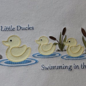 May include: Embroidered design of three yellow ducks swimming in a pond with green reeds. The text "Three Little Ducks Swimming in the Pond" is embroidered below the ducks.