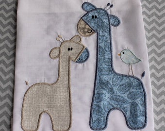May include: White fabric with appliqued giraffes and a bird. One giraffe is light tan, the other is blue with a leaf pattern. A small blue bird is perched on the blue giraffe's back. The design is outlined with a brown stitch.