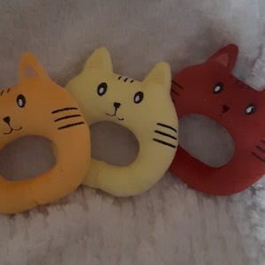 May include: Three plush cat-shaped rings in orange, yellow, and red. Each ring has a black embroidered face with eyes, a nose, and whiskers.