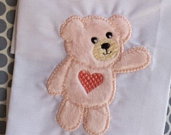 May include: A pink embroidered teddy bear with a red heart on its chest. The bear is standing with its right arm outstretched.