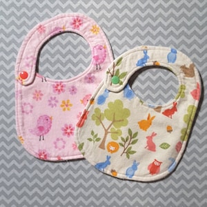 Op de afbeelding: Twee baby-slabbetjes, een roze met een bloemen- en vogelpatroon en een crème met een bosdierpatroon.